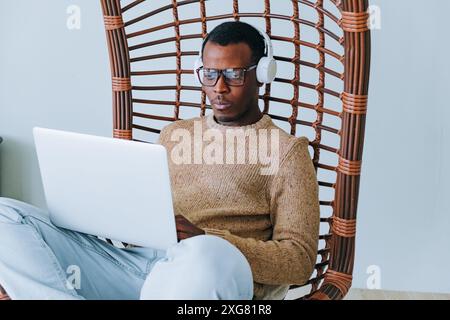 Ein afroamerikanischer Mann mit Kopfhörern und Brillen, der in einem gewebten Stuhl sitzt, konzentrierte sich auf die Arbeit am Laptop in gemütlicher Umgebung. Ein seriöser Black Guy-Programmierer ist leidenschaftlich für schwierige Aufgaben am Computer, Fernarbeit. Stockfoto
