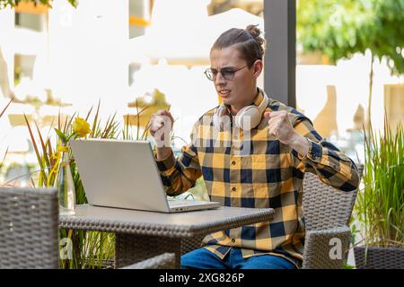 Gutaussehender junger Mann, der Laptop-Computer benutzt, um zu surfen, verliert überraschte plötzliche Lotterieergebnisse, schlechte Nachrichten, Verlust von Vermögen, scheitern. Guy sitzt draußen auf der Terrasse des städtischen Cafés. Lebensstil in der Stadt. Stockfoto