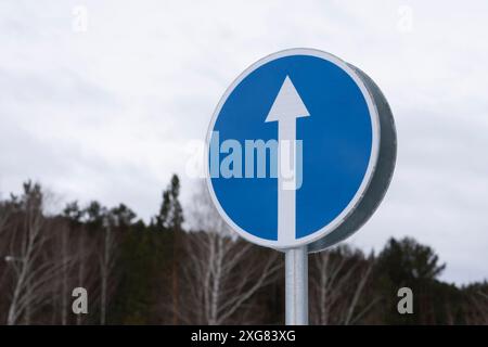 Ein blaues kreisförmiges Verkehrsschild zeigt einen weißen Pfeil, der nach oben zeigt und vor einer natürlichen Kulisse von Bäumen steht. Stockfoto