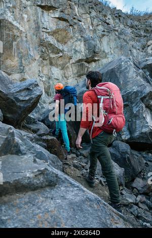 Zwei Wanderer mit Rucksäcken nähern sich einer felsigen Klippe und bereiten sich auf einen anspruchsvollen Aufstieg vor. Sie tragen Helme und Kletterausrüstung, bereit für den Outdoor-Einsatz Stockfoto