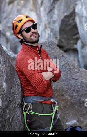 Ein selbstbewusster, sonnenverwöhnter Kletterer mit orangefarbenem Helm und roter Jacke liegt auf einem Felsen. Er lächelt mit überkreuzten Armen und zeigt seine Kletterausrüstung Stockfoto