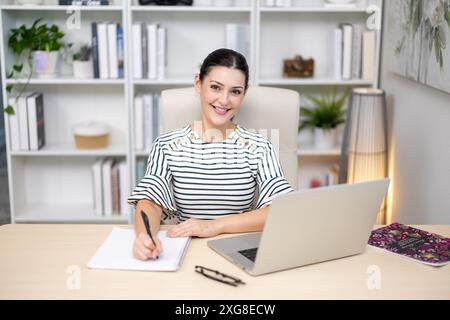 Ein brünettes weibliches Model, das im Büro am Computer arbeitet und ein gestreiftes Kleid trägt Stockfoto