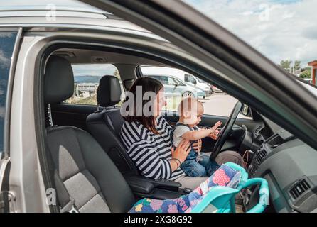 Mutter und Baby sitzen im geparkten Auto, während das Baby spielerisch das Lenkrad hält. Die Szene fängt den zärtlichen Familienmoment ein, betont Bindung, Erkundung, A Stockfoto