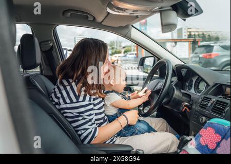 Mutter und Baby sitzen im geparkten Auto, während das Baby spielerisch das Lenkrad hält. Die Szene fängt den zärtlichen Familienmoment ein, betont Bindung, Erkundung, A Stockfoto