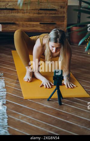 Mädchen Yoga Coach führt mit ihrer Gruppe einen Online-Kurs über Video Link mit Smartphone. Frau in gelber Activewear nimmt ihre Yoga-Session auf dem Smartphone auf und posiert auf der Matte in gemütlicher Atmosphäre Stockfoto