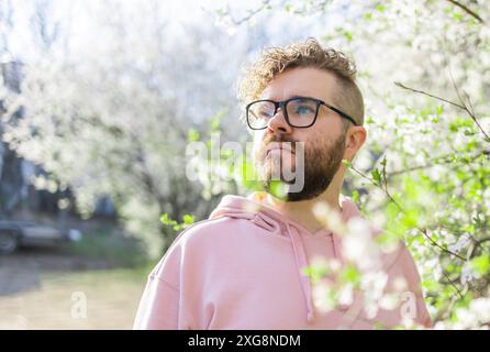 Mann mit Bart und Schnurrbart auf lächelndem Gesicht in der Nähe von Sakura-Blumen oder blühendem Frühlingsbaum. Weiches und sanftes Konzept. Bärtiger Mann mit stilvollem Haarschnitt Stockfoto