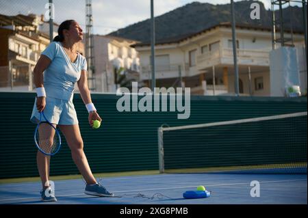 Frau spielt Tennis auf einem Platz im Freien, hält Schläger und Tennisball. Sonnenlicht verbessert die sportliche Szene. Netz, Zaun und Gebäude im Hinterhaus Stockfoto