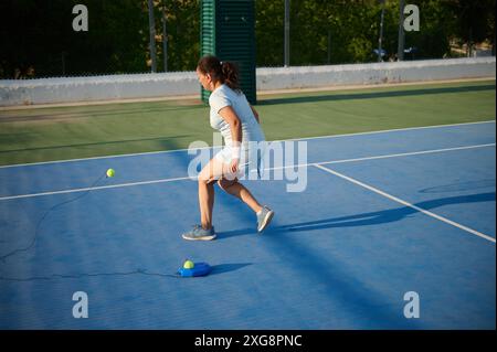 Frau in Sportkleidung, die Tennis auf einem Freiplatz mit einem Balltrainer übt. Ihre Fähigkeiten unter hellem Sonnenlicht zu perfektionieren. Stockfoto