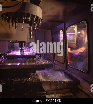 CNC-Fräsmaschine schneidet Metall mit Präzision, violette Funken fliegen, während der Bediener Prozesse durch Sicherheitseinhausungen in modernen Werkstätten überwacht. Stockfoto