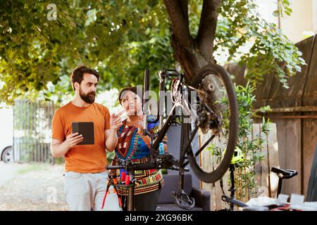 Engagiertes jugendliches interrassisches Paar mit digitalem Gerät zur Reparatur und Wartung moderner Fahrräder. Im Freien aktive lebhafte Mann und schwarze Frau, die das Internet für Fahrradwartung erkunden. Stockfoto