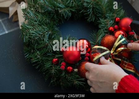 Frau, die dekoratives Band bindet Schritt für Schritt Anleitung zur Herstellung handgemachter Weihnachtskranz aus festlichen Dekorationen in roter Farbe. Weibliche Hände kreieren hausgemachte Kreationen. Neujahrsfeier. Winterferien Schritt 3 Stockfoto