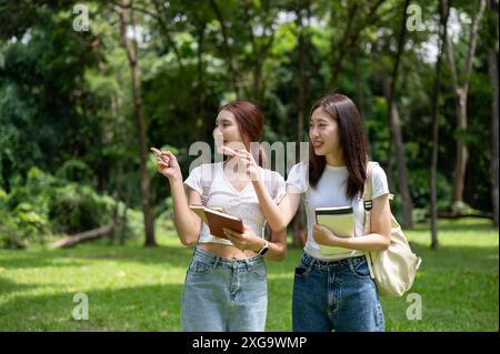 Zwei hübsche und glückliche junge asiatische Studentinnen mit Rucksäcken zeigen mit dem Finger auf etwas, reden und laufen im grünen Park Stockfoto