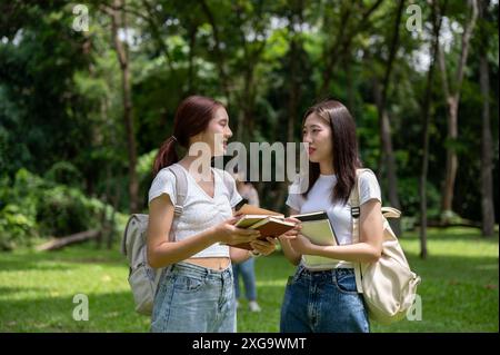 Zwei hübsche und glückliche junge asiatische Studentinnen mit Rucksäcken unterhalten sich beim gemeinsamen Spaziergang im Campus-Park. Freundschaft und Erziehung Stockfoto