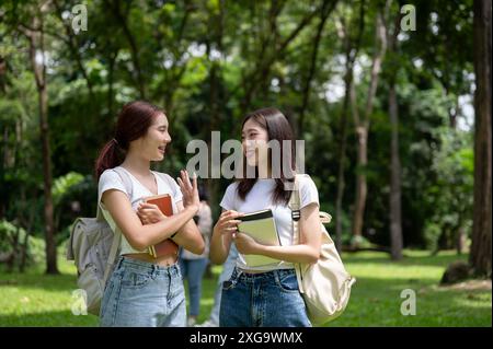 Zwei hübsche und glückliche junge asiatische Studentinnen mit Rucksäcken unterhalten sich beim gemeinsamen Spaziergang im Campus-Park. Freundschaft und Erziehung Stockfoto