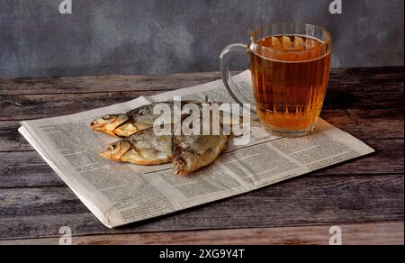 Drei große, getrocknete Flussfische und ein Becher mit leichtem Bier auf einer alten Zeitung und auf einem Holztisch. Nahaufnahme. Stockfoto