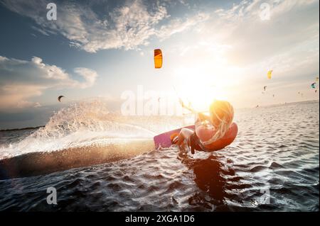 Professionelle Kitesurferin reitet mit einer Planke in den Händen auf einem See mit Meerwasser bei Sonnenuntergang. Wasserspritzer und Sonnenstrahlen. Stockfoto