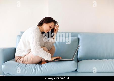 Junge nachdenkliche Frau in Freizeitkleidung, die mit ihren Beinen auf dem Sofa sitzt, die Tastatur auf dem Laptop benutzt, auf den Computerbildschirm starrt und nach Wichtigem sucht Stockfoto