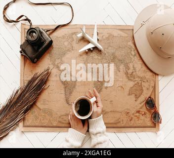 Draufsicht auf weibliche Hände, die eine Tasse schwarzen Kaffee halten, über einer Vintage-Weltkarte mit Reisezubehör, Retro-Kamera und darauf liegendem Spielzeugflugzeug Stockfoto