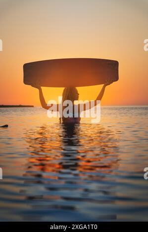 Attraktive junge Frau in einem Taucheranzug, die hüfttief im Meerwasser steht und ein Kitesurfbrett auf dem Kopf hält, das antike ägyptische Motive zeigt Stockfoto