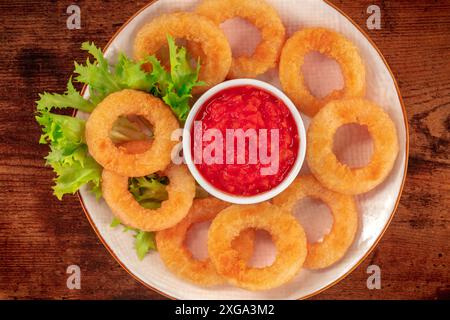 Kalmaringe aus nächster Nähe auf einem Holzhintergrund. Frittierte Calamari-Ringe mit grünem Salat und würziger Tomatensauce, darüber gebratener Schuss Stockfoto