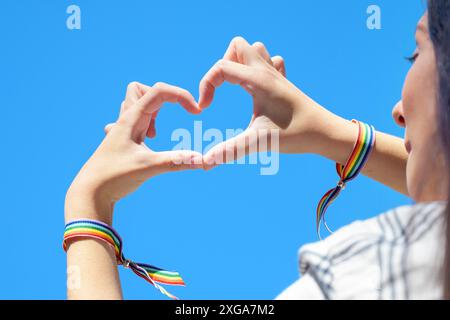 Nahaufnahme von Frauenhänden in einem Regenbogenarmband, die eine Herzform auf blauem Himmel bilden. Hochwertige Fotos Stockfoto