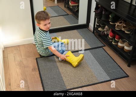 Ein Kind zieht Schuhe an, die im Flur stehen. Das Baby zieht gelbe Stiefel an, um spazieren zu gehen. Kind im Alter von 3 Jahren (dreijähriger Junge) Stockfoto