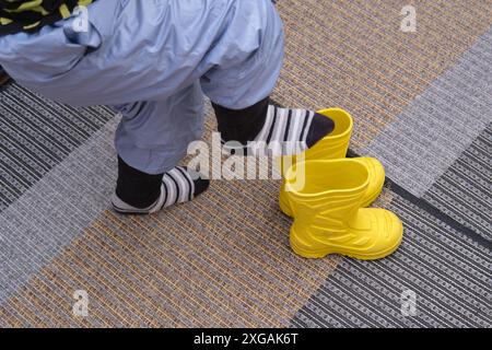 Ein Kind zieht Schuhe an, die im Flur stehen. Das Baby zieht gelbe Stiefel an, um spazieren zu gehen. Kind im Alter von 3 Jahren (dreijähriger Junge) Stockfoto