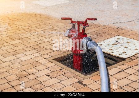 Roter Hydrant mit angeschlossenem Schlauch mit Spritzwasser für die Brandbekämpfung auf dem Gehweg im Sommer Stockfoto