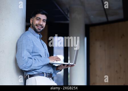 Lächelnder Mann mit Laptop, während er sich an die Säule lehnt, im modernen Büro, im Kopierraum Stockfoto