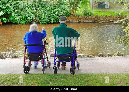 Senioren reifes Rentneralter Paar Rückansicht ruht neben dem Dawlish Wasserstrom sitzend auf Mobilitätshilfen am kühlen Juni Tag Devon England UK Stockfoto