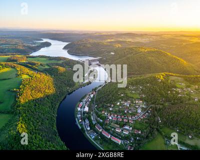 Ein Luftbild des Orlik Reservoir in Tschechien bei Sonnenuntergang zeigt einen gewundenen Fluss, Hügel und eine malerische Stadt im Tal mit leuchtendem blauem und goldenem Himmel. Stockfoto