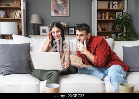 Einfaches Leben Junge Verlobte Paare sitzen zu Hause, verbringen wertvolle Zeit, Frau auf der Suche nach Hochzeitskleid auf Laptop, Mann liest ein Buch, organisiert Stockfoto