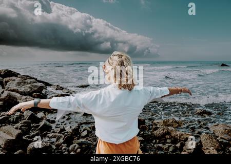 Eine ältere Frau mit weißem Hemd streckt sich mit offenen Armen, mit Blick auf das Meer unter einem bewölkten, stürmischen Himmel, stehend an einem felsigen Ufer. Stockfoto