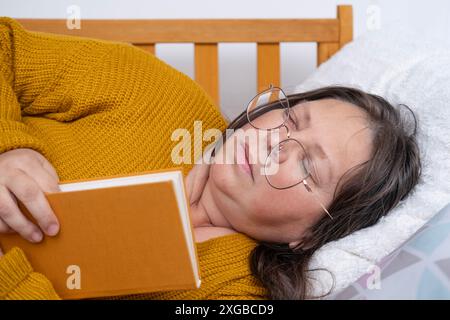 Frau 50 Jahre alt in gelbem Pullover und Brille Buch lesen und eingeschlafen, Work-Life-Balance, Müdigkeit, Müdigkeit oder Erschöpfung, Lesen kann heller werden Stockfoto