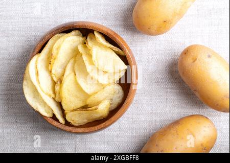 Gesalzene Kartoffelchips in einer Holzschale auf Leinengewebe. Chips, dünne Scheiben frittierter Kartoffeln und auf den richtigen rohen Kartoffeln. Stockfoto