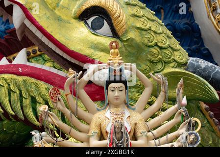 Detaillierte Statue einer mehrarmigen Gottheit mit verschiedenen Symbolen und einem goldenen Buddha, vor einem farbenfrohen Drachenhintergrund in einem Koh Samui templ Stockfoto