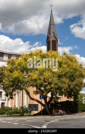 Dunkler Turm der evangelischen Kirche am Ufer von Vah. Teil der Innenstadt. Großer blühender Baum vorne. Piestany, Slowakei. Stockfoto