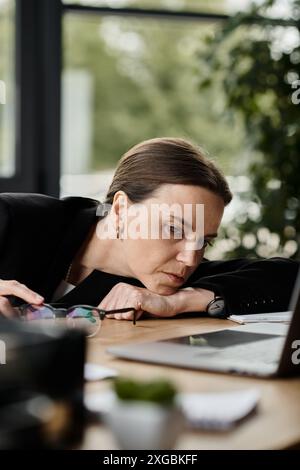 Eine Frau mittleren Alters, die Stress erlebt, wird in einem Büro über ihren Laptop gebeugt. Stockfoto