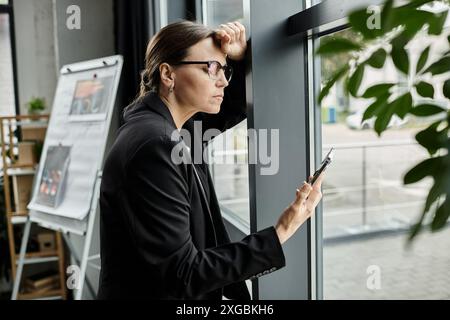 Eine Frau mittleren Alters in Brille lehnt sich gegen das Fenster und blickt auf den Bildschirm des Telefons. Stockfoto