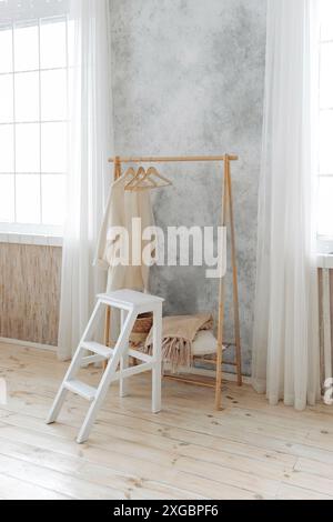 Stilvolle Holzgarderobe und weißer Holzleiterhocker in einem geräumigen Innenraum mit großen Fenstern mit Vorhängen. Niemand. Holzboden im Apartment Stockfoto