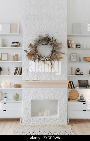 Weiße Regale mit Büchern und Dekor in der Nähe von Kamin mit Kranz aus getrockneten Blumen in einem modernen, stilvollen Wohnzimmer im skandinavischen Stil. Niemand Stockfoto