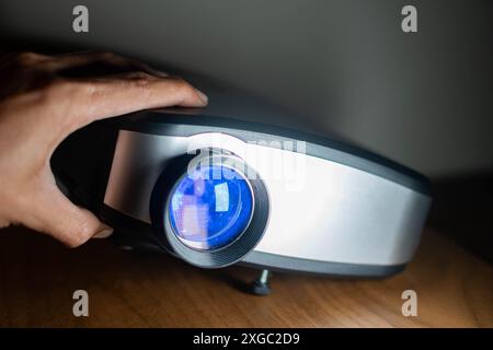Eine Nahaufnahme der Hand einer Person, die einen modernen Digitalprojektor auf einer Holzoberfläche anpasst. Das Objektiv des Projektors wird mit blauem Licht beleuchtet. Stockfoto