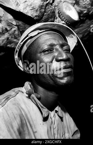 Ein vertikales Graustufenporträt eines afrikanischen Arbeiters mit Schutzhelm und Stirnlampe in unterirdischem Platinum-Palladium-Bergbau und -Maschinen Stockfoto