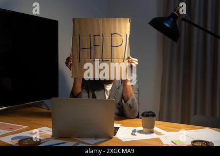 Junger Geschäftsmann, Der Pappe Mit Hilfetext Am Arbeitsplatz Hält. Junger asiatischer Geschäftsmann, der mit Stress am Laptop-Schreibtisch mit Pappe arbeitet Stockfoto