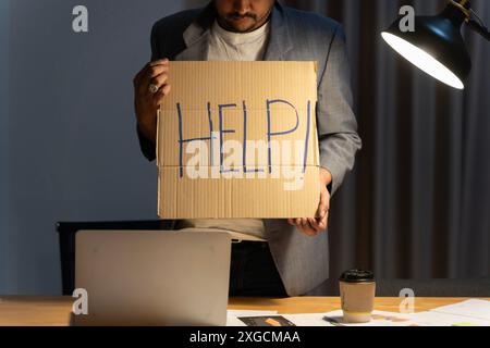 Junger Geschäftsmann, der an einem Computer-Laptop arbeitet und um Hilfe bittet, ein Pappschild hält, das verzweifelt und depressiv im Stress ist überwältigt. Überarbeitet Stockfoto