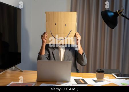 Ein junger Geschäftsmann hält einen Karton mit einem Smiley-Gesicht vor seinem Kopf, mit einer Pappschachtel auf dem Kopf, mit einem Smiley-Gesicht. Stockfoto