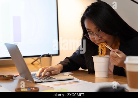 Asiatische unabhängige, intelligente Geschäftsfrau isst Instant-Nudeln, während sie nachts im Wohnzimmer am Laptop arbeitet, glückliches asiatisches Mädchen sitzt auf dem Schreibtisch Stockfoto