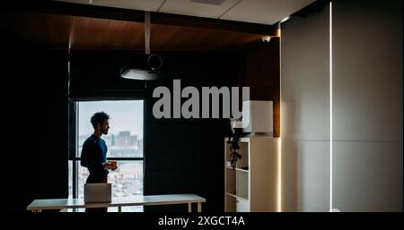 Guten Morgen dieses jungen, hübschen Designers beginnt mit Kaffee und positiven Gedanken, wenn er von seinem Büro aus die wunderschöne Stadtlandschaft ansieht Stockfoto