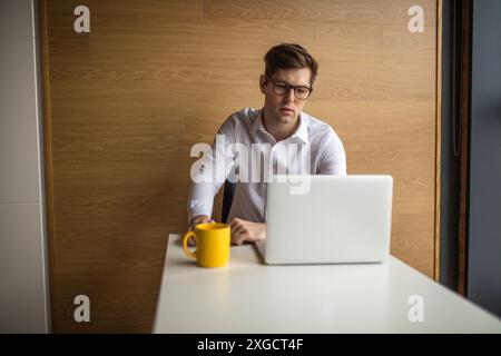 Konzentrierter europäischer junger Angestellter in Brille, der mit nachdenklichem Ausdruck auf den Laptop-Bildschirm schaut, über seinen Geschäftsbericht nachdenkt, Papa macht Stockfoto