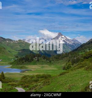 Panorama mit Parkbank auf einem Wanderweg mit Blick auf einen See und die umliegenden Berge im Arlberg, schneebedeckter Wolkengipfel Stockfoto